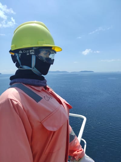 The image, oriented correctly, depicts a person in protective gear on a vessel at sea, facing towards the right. The person is wearing a bright yellow hard hat, clear protective goggles that obscure their face, and a dark blue neck gaiter covering their lower face and neck. They are dressed in a reddish-orange long-sleeved coverall or work uniform, which features a reflective grey stripe on the right shoulder/sleeve and some faint dark text (possibly "SG" or "50") on the right chest area. A white work glove is partially visible on their right hand, which appears to be holding onto a white metal railing, suggesting they are on a boat or ship.

The background shows a wide expanse of deep blue sea with gentle ripples, extending to a distant horizon where faint landmasses or mountains are visible. The sky is clear, light blue with sparse, wispy white clouds. The bright, even lighting indicates it is daytime, likely morning or afternoon. The overall scene suggests a work environment on the water, possibly in a maritime or offshore setting. The location context provided is Kunak, Malaysia, which is a coastal area. No emotions are discernible due to the protective gear. Visible text on the goggles strap includes "brI g" and a triangular logo.