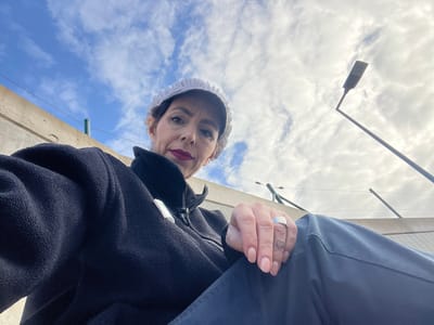 A low-angle photograph shows a person in Roodt-sur-Syre, Luxembourg, facing the camera. The person wears a white lace bonnet and a dark fleece jacket, with blue trousers visible above. Their left hand, adorned with a ring and a heart tattoo on the finger, is raised. Behind them, the sky is visible with scattered clouds. A streetlight pole extends into the frame from the lower left. A beige concrete structure is seen partially in the background.