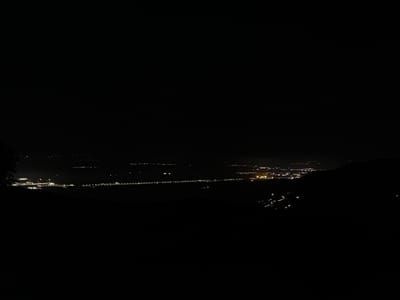 The image presents a broad, dark nighttime landscape dominated by the distant lights of a town or city, viewed from a high vantage point, likely a hill or mountain. The upper half of the frame is almost entirely black, signifying the night sky, with no stars or moon visible.
The main subjects are the numerous artificial lights scattered across the lower half of the image. These lights appear as small, luminous specks, varying in brightness and color, predominantly yellow and white. They form distinct patterns, outlining the extent of human settlement. A prominent linear cluster of lights stretches horizontally across the mid-ground, possibly indicating a major road, railway, or an illuminated feature like a riverbank. To the left, a more concentrated and brighter area of lights suggests a bustling part of the town or a specific facility. On the right, groups of lights ascend gently, hinting at development on sloping terrain.
The foreground is cloaked in deep shadow, with only faint, indistinct dark shapes suggesting the presence of foliage or uneven terrain, from which the photograph was taken. Similarly, the areas beyond the illuminated settlement are submerged in darkness, with subtle dark outlines in the far distance indicating further hills or mountains.
The time of day is clearly night. The air appears mostly clear, allowing the distant lights to be seen distinctly, though there is a slight atmospheric haze. No specific activity or event is discernible beyond the static display of urban illumination. No people or identifiable objects other than the lights and the dark landscape are visible. There is no visible text. The scene conveys a sense of peaceful vastness and the quiet glow of civilization against the natural darkness. Given the context, this is a night view of Galabovo, Bulgaria, or its immediate surroundings, from an elevated perspective.