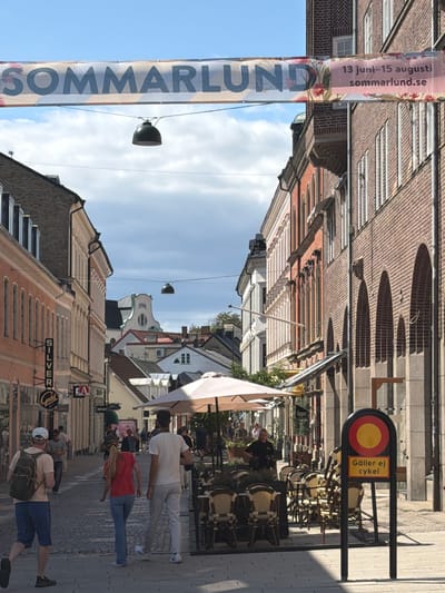 The image depicts a street scene in Lund, Sweden, likely during the summer festival "Sommarlund," as indicated by a banner across the street that reads "SOMMARLUND." The banner also includes dates "13 juni - 15 augusti" and a website "sommarlund.se." The street is paved with cobblestones and lined with buildings. On the left, there are buildings with light-colored facades and awnings, and signage for "SILVER" and "Guild." Further down the street, more buildings are visible, including one with a white facade and decorative elements. On the right side of the street, buildings are constructed of brick, featuring arched ground-floor openings. A sidewalk cafe with tables and chairs is set up on the right, covered by a large white umbrella. In the foreground, three people are walking away from the camera. A man with a backpack is on the left, followed by a woman in a red top and blue jeans, and a man in a white t-shirt and light-colored pants. A street sign with a red circle and yellow circle, and text "Gäller ej cykel" (Not applicable to bicycles) is prominently displayed on the right side of the image. The sky is blue with some clouds. Various shops and establishments are indicated by signage along the street.
