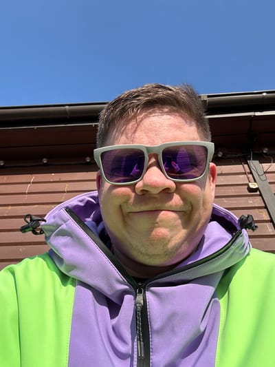 A close-up, head-and-shoulders image of a male subject, captured in Vasil'evo, Russia, under clear blue skies. The subject, with short brown hair and a fair complexion, exhibits a slight smirk. He wears light gray-framed sunglasses with purple reflective lenses. A smartphone, held by a hand with light-colored fingernails, is clearly reflected in both lenses, indicating a self-portrait. The subject is dressed in a two-tone outdoor jacket; the right side (viewer's left) is neon green, and the left side (viewer's right) and the hood are lavender. The jacket features a visible black zipper and a drawstring with an adjuster on the hood. The background consists of the upper portion of a brown wooden building with horizontal siding panels, above which a dark-colored gutter and roofline are visible. Several small, round, light-colored fixtures are strung horizontally across the upper section of the wooden wall.