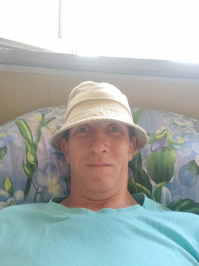 A close-up, selfie-style photo shows a person wearing a light-colored bucket hat and a light blue t-shirt. The person is lying down on a pillow with a blue floral pattern. The background consists of a wall paneled with light-colored wood and a bright window at the top. The lighting is natural and appears to be coming from the window, creating a bright, slightly washed-out effect. The person's face is visible, and they are looking directly at the camera. The image does not provide specific details about the location within Соколово, Russia, beyond suggesting an indoor setting, possibly a home.