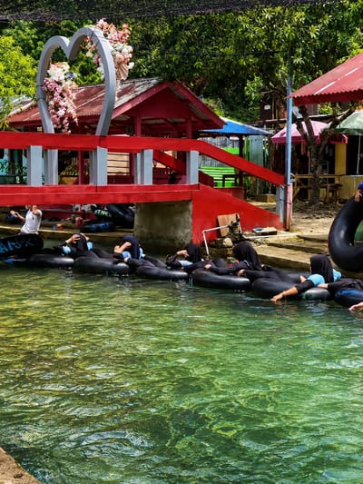 In the city of Silaiyajae, Indonesia, a group of people are seen floating on inner tubes in a clear, greenish body of water. The water appears shallow, with sunlight filtering through to illuminate the textured bottom. A red wooden bridge with grey accents crosses over the water, adorned with a large, grey heart sculpture decorated with pink and white flowers. Beyond the bridge, several small red buildings with corrugated tin roofs are visible, nestled amongst lush green trees. The overall atmosphere suggests a recreational area or a tourist spot.