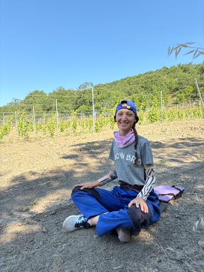 A smiling woman sits cross-legged on the ground in a vineyard. She is wearing a blue baseball cap, a grey t-shirt with "HOW BRO" printed on it, a pink neck gaiter, and blue overalls. She has on arm sleeves with a pattern and is wearing athletic shoes. Her left hand rests on her knee, and her right hand is on the ground. Behind her, rows of young grapevines are planted in cultivated soil, supported by metal posts and wires. The vineyard is situated on a hillside covered with green trees, under a clear blue sky. The ground is dry and dusty, with some dry grass and shadows cast from unseen foliage.