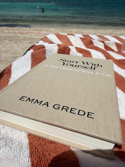An eye-level, medium shot captures a book resting on a striped beach towel with an orange and white diamond pattern. The book, titled "Start With Yourself: A New Vision for Work & Life" by EMMA GREDE, is positioned on the right side of the frame, with its cover facing upwards. The towel is laid out on a sandy beach, with the turquoise ocean visible in the background. Two figures are seen swimming in the water. The lighting suggests a sunny day.