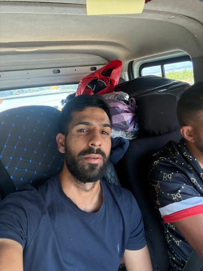 Two men are seated in the interior of a vehicle, likely a van, as indicated by the overhead storage compartments and visible car ceiling. The man in the foreground is on the left, facing the camera, with dark hair and a beard. He is wearing a navy blue t-shirt. To his right, partially visible, is another man wearing a black t-shirt with a gold patterned design and a red and white striped trim on the sleeve. Luggage, including a red bag and at least one large black suitcase, is visible in the background, indicating travel. The seats appear to be upholstered with a dark fabric featuring a geometric blue pattern. The environment outside the vehicle's window suggests a sunny day, with trees and a distant landscape visible. The location is stated to be Gara Elin Pelin, Bulgaria.