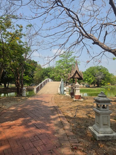 The scene depicts a park in Phra Nakhon Si Ayutthaya City Municipality, Thailand. A brick walkway leads towards a small Thai-style pavilion on a pond. Stone lanterns line the walkway. Trees are on either side, some with bare branches. The sky is partially visible through the branches.