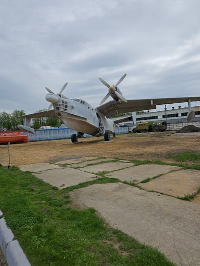 The image depicts an Antonov An-26 aircraft on display. The aircraft is painted in a light grey color scheme, with propellers featuring yellow tips. It is parked on a concrete slab surrounded by a grassy area. In the background, there is a building with multiple windows and a military vehicle visible. The sky is overcast. The image seems to be taken at the Vadim Zadorozhny's Museum of Technology in Arkhangel’skoye, Russia.