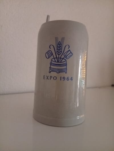 This is a medium shot photograph of a ceramic beer stein. The stein is light grey with a blue design on its front. The design features a crest with wheat stalks in the center, flanked by two tankards on either side. Below the crest, the words "EXPO 1964" are printed in blue. The stein has a metallic lid with a small handle. The stein is positioned on a white, flat surface, possibly a table or shelf, with a white textured wall in the background. The lighting suggests an indoor setting, possibly during the day. The image is slightly blurry, and the focus is on the stein. The context provided indicates the stein is related to Langenthal, Switzerland, and the year 1964, likely commemorating an event such as a trade fair or exhibition held in that location. No people are visible in the image.