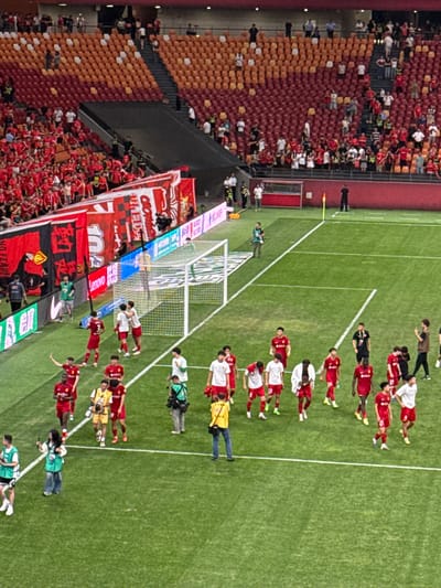 An overhead perspective shows a soccer field in Longxing, China, with stadium seating on the left and players aligned on the field. A soccer goal is present. A crowd gathers in the stands, displaying banners with Chinese characters. Some individuals in the stands wear red, while the seats are orange and red. A red line divides the seated fans. The soccer players are lined up facing the fans. Photographers and personnel surround the soccer players. The field is green with white lines.