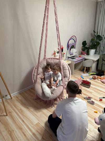 The image shows an indoor scene, seemingly a child's playroom, potentially located in a residential setting rather than the city of Krapivka, Russia, as the location cannot be confirmed from visual information. Two young children are sitting together in a pink macrame hanging chair; one child is lying down. A man in a white t-shirt and dark pants is kneeling and facing the chair, likely interacting with the children. The room has light-colored wood flooring and light-colored walls. There are various toys and items scattered across the floor, including shoes, a toy animal, and other playthings. A plant is visible near a desk with children's art supplies.