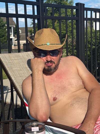 A Caucasian man with a beard and a tan cowboy hat with a turquoise embellishment is sitting in a lounge chair outdoors, leaning on his hand and wearing sunglasses. He is shirtless, exposing his chest and arms, and appears to have a sunburn on his chest. He is wearing pink patterned shorts. A metal fence with vertical slats is visible behind him, with trees and houses in the background. A tumbler cup is in the foreground on the chair. The setting is likely a residential backyard or poolside area in Allen Township, United States of America.
