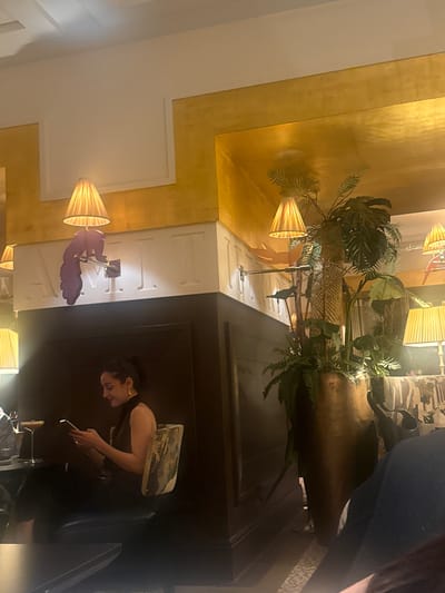 A sideways-oriented interior scene shows a woman sitting in what appears to be the lobby or lounge of a hotel or upscale establishment, potentially in Bellevue, Switzerland. The room features a combination of golden and white wall finishes, decorative lamps with pleated shades, and large potted plants. The woman is seen looking at a mobile phone, dressed in dark attire, with what looks like a patterned bag or item placed behind her. The atmosphere suggests a stylish, possibly luxurious setting.