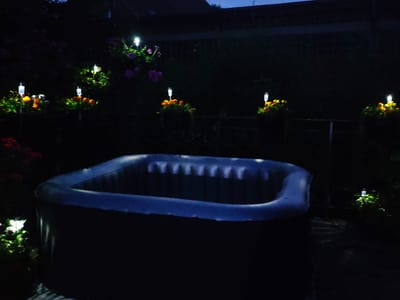 The image depicts an inflatable hot tub situated in a dark outdoor environment, likely a backyard, at night. The hot tub is a prominent, dark-colored object in the foreground, with its inflatable rim visible. Several small, solar-powered garden lights are scattered throughout the scene, illuminating clusters of orange and pink flowers, as well as foliage. The lights create a gentle glow, highlighting the plants against the darkness. The background is mostly obscured by shadows, with hints of structures and more vegetation. There are no people visible in the image, and no specific actions or interactions are taking place. The overall impression is one of a quiet, illuminated garden scene at night, with the hot tub as a central feature. The location is stated to be in the city of Lügde, Germany.