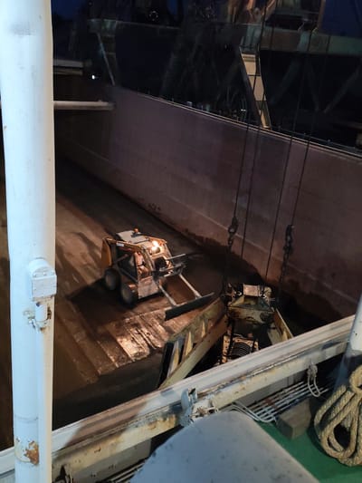 The image shows the interior of a ship's cargo hold at night, illuminated by artificial lights. A small orange and black CASE skid steer loader is positioned on the dark, wet floor of the hold, with its front-mounted bucket lowered. The loader appears to be cleaning or clearing the hold. Above and to the right of the loader, a large industrial grab or bucket suspended by heavy chains is visible, suggesting ongoing cargo operations. The cargo hold walls are tall and dark, with some reflections on the wet floor. A thick white pipe runs vertically along the left side of the frame, obscuring part of the scene. In the foreground, a portion of the ship's deck is visible, including a thick coiled rope and some metal railings. The scene is industrial and appears to be operational. The location provided, Chelganovo, Russia, is a rural settlement and not typically associated with large cargo ships.