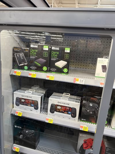 This image captures the interior of a locked retail display case, likely within a store in Worcester, United States. The cabinet is well-lit and houses various gaming accessories, predominantly external storage devices and controllers.

On the left side of the display, several external hard drives are arranged vertically. Two boxes for "Seagate Game Drive for Xbox" are prominent, with one indicating "5TB HDD" and showing a white drive, and the one below it marked "2TB HDD" displaying a dark-colored drive. Below these is a "WD BLACK P10" portable game drive. Associated price tags read "$59.00," "$85.00," and "$124.00" respectively, with some labeled as "Rollback."

The right side of the cabinet is dedicated to gaming controllers. A black "XBOX FUSION" controller box is visible on an upper shelf, priced at "$69.00." Below it are two "BACKBONE" mobile gaming controllers, designed to attach to a smartphone. The packaging clearly depicts a phone screen showing "Call of Duty: Mobile." Both Backbone controllers are priced at "$99.00." On the lowest shelf, an "XBOX ELITE SERIES 2" controller box is partially visible, with its price tag suggesting "$179.00."

The background beyond the items is a perforated metal panel, and reflections on the glass door indicate bright overhead fluorescent lighting, typical of a retail setting. The visible text also includes "GAME PASS" and various Xbox logos, confirming the gaming theme. No people or specific activities are visible, just the displayed merchandise.
