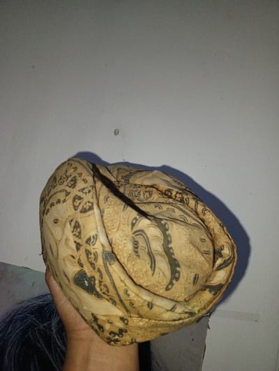 A human hand holds a folded textile item, possibly a headdress or cap, patterned with dark, intricate designs against a light tan or beige background. The textile appears to be made from a woven fabric, exhibiting folds and a somewhat irregular, rounded shape. The patterns include stylized floral or abstract motifs. The hand, positioned in the lower left quadrant, holds the item from below. The background consists of a plain, light-colored wall, likely painted off-white or light gray, showing minor imperfections and possibly some dust or small marks. A distinct shadow from the textile item is cast onto the wall, indicating a light source from the upper right or front. There is no visual information within the image to identify the location as Na Sang, Laos, or any other specific geographical area.