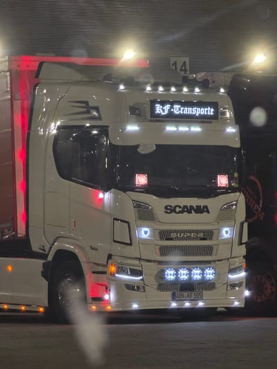 The image shows a white Scania semi-truck parked at night. The truck has a license plate that reads "ARN KF 505". The truck is decorated with red and white lights. In the background, there is a building with horizontal lines and a sign with the number "14". There is also another truck visible to the right of the main truck, with some lettering on it that is partially obscured. The overall scene suggests the truck is at a loading dock or a parking area.