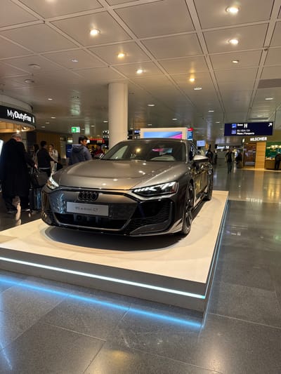 The scene depicts an interior view of a commercial area within Munich Airport, located in Oberding, Germany. A grey Audi RS e-tron GT electric vehicle is centrally displayed on a raised, rectangular white platform, which features a luminous blue LED strip along its base. The car faces slightly towards the left of the viewer, with its headlights illuminated.
The immediate environment consists of a dark grey tiled floor and a white grid-paneled ceiling equipped with multiple recessed circular light fixtures. White support columns are interspersed throughout the area.
In the left background, a "MyDutyFree" sign is visible above a cluster of individuals. One person, dressed in a dark coat, is partially visible from behind, appearing to attend to luggage. Other blurred figures are present in this section.
In the right background, overhead signage indicates "Gate H" and "EU / EEA / CH Pässe / Paspoorten" (Passports), along with a "HILSCHER" store sign below. Several individuals are seen walking or standing in this area, consistent with general airport passenger traffic. Digital display screens are also faintly visible further in the background.