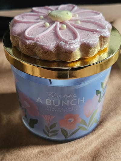 A close-up shot reveals a "Thanks A Bunch Rose Bouquet Scented Candle" with a cake-like lid wrapped in plastic. The lid features a pink floral design with a yellow center and white accents. The candle is positioned on a brown textured surface. The location is unknown.