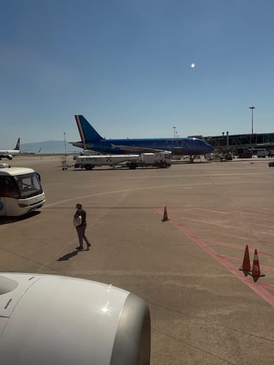 The image shows an airport tarmac in Cinisi, Italy, with an ITA Airways Airbus A320 parked at a gate. A fuel truck is positioned near the plane's wing. There are two orange cones on the tarmac. In the foreground, part of an aircraft engine is visible. A man is walking near the plane, carrying a white bag. Another aircraft, with a Ryanair logo on its tail, is parked farther away. In the distance, mountains can be seen. The sky is clear.