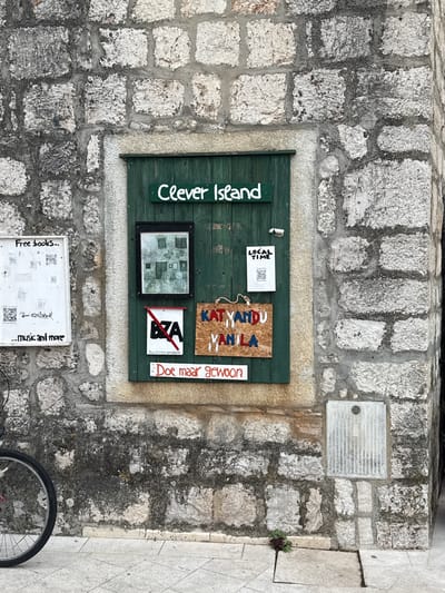 The image shows a section of a rough stone wall in the city of Grad Stari Grad, Croatia. Mounted on the wall is a dark green wooden door or panel with various signs and artwork displayed on it.

At the top of the green panel, a white sign reads "Clever Island" in white lettering. Below this, a framed black and white drawing depicts several figures standing in front of buildings. To the right of the drawing is a smaller white sign with black lettering that says "LOCAL TIME" and includes a QR code.

Further down, to the left of the drawing, is a white sign with a red diagonal line through the letters "BZA" and the text "Busy Zombie Apocalypse" below. To the right of this, a wooden sign with a rough texture has "KATYANDU YANILA" written on it in colorful letters.

At the bottom of the green panel, a white sign with red lettering reads "Doe maar gewoon".

To the left of the green panel, a white rectangular sign is mounted on the stone wall. It has the text "Free books..." at the top and "...music and more" at the bottom, with several QR codes interspersed.

In the foreground, the lower left corner of the image shows a portion of a bicycle wheel. The ground is paved with light-colored stone tiles. A small electrical box with vertical slats is mounted on the stone wall on the right side of the image. A small plant is growing from a gap in the paving stones near the bottom center of the image. The overall scene suggests an outdoor display or bulletin board in a historic town setting.