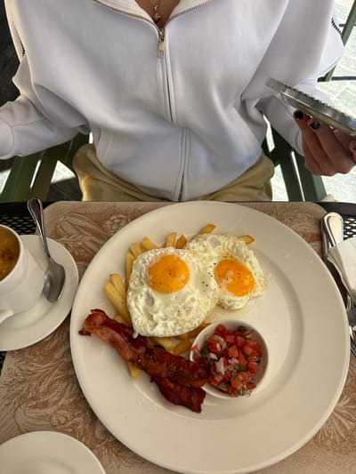 A person is seated at an outdoor table, about to enjoy a breakfast. A white plate holds two sunny-side-up eggs, a small bowl of diced tomatoes, and two strips of bacon, all served on a bed of French fries. Utensils are placed around the plate. A cup of coffee sits on a saucer nearby. The person is wearing a white zippered hoodie.