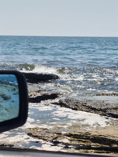 The image depicts a rocky shoreline with the Caspian Sea in the background. The water is calm with gentle waves lapping against the shore. Sunlight glints off the water's surface, creating a sparkling effect. In the foreground, a side-view mirror of a vehicle is visible on the left, reflecting the sea and the rocky terrain. A small bird is foraging near the water's edge among the rocks. The sky is clear and bright, suggesting a sunny day. The scene is set in Saiyn, Kazakhstan, on the shores of the Caspian Sea.