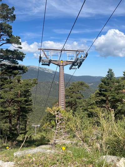 The image depicts a ski lift structure situated within a mountainous landscape near Real Sitio de San Ildefonso, Spain. The lift structure is composed of a metal tower with a ladder extending along its length, supporting cables that stretch into the distance and connect to other lift towers, one of which is partly visible. The environment includes a clear blue sky with scattered clouds. The surrounding terrain is forested with diverse vegetation, including coniferous trees and grassy areas with yellow wildflowers. A few peaks can be seen in the background.