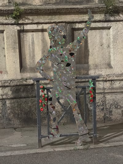 The image depicts a silhouette of a person, constructed from numerous pieces of broken CDs, standing in front of a stone wall in Vienne, France. The silhouette is positioned outdoors, on a paved surface that appears to be a sidewalk adjacent to a street. A dark metal railing is situated in front of the silhouette, with a string of red artificial flowers hanging from it on the left side and a similar arrangement on the right. The CD mosaic reflects light, creating a shimmering effect. The stone wall behind the sculpture is weathered and has some vegetation growing from its upper ledge. The lighting suggests it is evening or nighttime, with shadows cast on the ground.