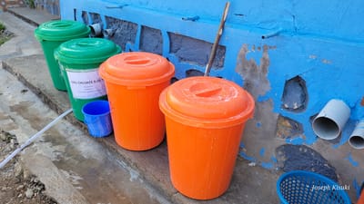 This outdoor image captures a water collection or hygiene station, likely in Bunia, Democratic Republic of the Congo, given the provided context. The scene is dominated by several plastic containers, two bright green and two vibrant orange, with matching lids, resting on a concrete ledge. A light blue wall, with some flaking paint revealing underlying grey plaster and natural stone, forms the background. Several grey and blue pipes are integrated into or extend from this wall, suggesting a water distribution system.
On the middle green container, a white label is visible with French text. The OCR reads: "EAU CHLORÉE 0,05 %" (Chlorinated water 0.05%), "Ne pas boire" (Do not drink), "Préparée le :" (Prepared on:), and "À renouveler toutes les 24 heures" (To be renewed every 24 hours). This indicates the green containers hold treated water, likely for sanitation purposes (e.g., handwashing) rather than consumption, requiring regular replenishment.
A small blue plastic bucket sits near the labeled green bin, and a white hose lies across the concrete ground, disappearing behind the bins and extending towards the left foreground, where the ground appears wet, suggesting water use or spillage. A wooden stick leans against the blue wall between the orange bins and the pipes. To the bottom right, a blue plastic basket is partially visible. The bright lighting suggests it is daytime. The overall impression is a functional, rudimentary, but essential water point. A watermark "Joseph Kisuki" is present in the bottom right corner.