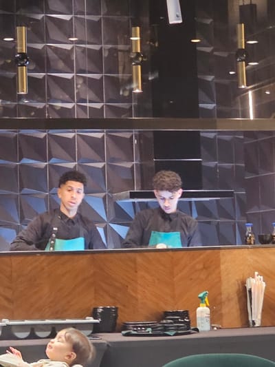 A mirrored bar reflects a young man in a black shirt and blue apron. The mirrored wall has geometric patterns and gold light fixtures. On the actual bar, behind the man in the reflection, are straws, a spray bottle, and stacks of plates. A child's face is visible near the edge of the bar. The overall tone suggests a modern and reflective atmosphere.