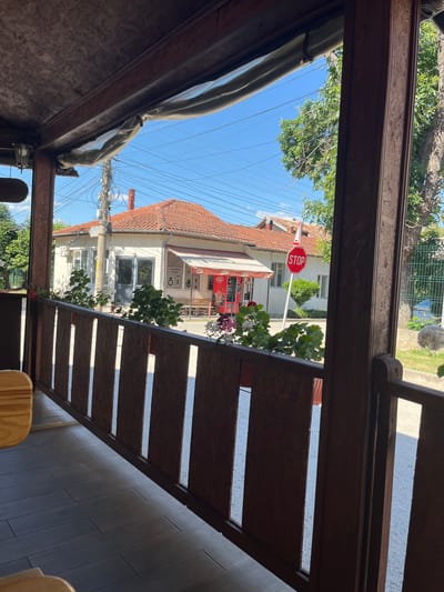 This is a photograph taken outdoors, looking through a wooden-framed opening onto a street scene. The image is oriented vertically, with the sky and trees appearing at the top, and the street and buildings below.
The scene appears to be a quiet street in Dulovo, Bulgaria, on a clear, sunny day, suggesting midday. The dominant subjects are buildings with red-tiled roofs and white facades, typical of European architecture. A prominent object in the scene is a red octagonal STOP sign, indicating a road intersection. There are also trees with green foliage, some power lines crisscrossing the sky, and a metal fence in the background. In the foreground, a wooden railing with vertical slats partially obscures the view.
The overall impression is one of a peaceful, ordinary day in a small town. There is no discernible activity or event taking place; it's a snapshot of a static street scene. Notable details include the vibrant blue sky, the sunlight casting shadows, and the tiled roofs of the houses.
There is visible text on the STOP sign. Additionally, on one of the buildings, there is text that appears to be a business name or sign, with what looks like an "$" symbol and the Cyrillic letters "ЗАКАНА". Another sign, possibly for a shop, has the text "BAIKAL". A red vending machine is also visible on one of the buildings with text in Cyrillic.