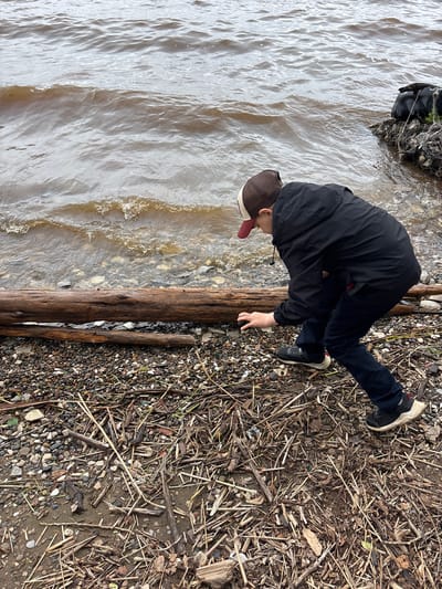 The image displays a male youth bent over at the water's edge in Kryukovo, Russia. The individual wears a brown and beige baseball cap, a black hooded jacket, dark trousers, and black sneakers, with their right hand extended towards the ground as if examining or picking up an object. The shoreline consists of varied-sized pebbles, gravel, and a dense accumulation of natural debris, including dry sticks, reeds, and dried leaves. Two prominent, weathered driftwood logs lie horizontally near the water, with the larger log partially submerged. The water itself is murky, grey-brown, exhibiting small ripples and minor wave action. In the upper right background, a dark, potentially plastic or fabric, bag-like object rests on a rocky bank at the water's periphery, partially entangled with what appears to be netting or wire. The outdoor scene is illuminated by diffused, overcast light.
