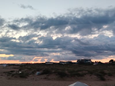 The image shows a beach and a distant treeline with buildings, set against a cloudy sky during sunset. The foreground consists of sand dunes with sparse vegetation. Several pieces of debris, including driftwood and plastic, are scattered on the sand. In the middle ground, there are several low-rise buildings, some with modern architecture, and trees. The sky is filled with dark, dramatic clouds that are lit from below by the orange and pink hues of the setting sun. The location is Blagoveshchenskaya, Russia. No people or specific actions are visible in the image.