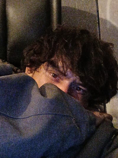 A person peers from behind a grey hoodie, only their eyes and part of their nose visible. They have dark, curly hair that frames their face. The background is blurred and appears to be a dark wall. The image is grainy. The person is indoors.