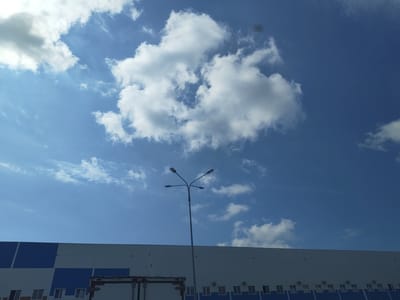 The image captures a view of a modern industrial building with a facade of white and blue panels, located in Prikubanskii, Russia. A large white semi-trailer truck is partially visible in the foreground. Above the building, a bright blue sky is filled with scattered white cumulus clouds. A tall lamppost with four lights is prominently positioned in the center of the frame, silhouetted against the sky. The overall scene suggests an outdoor industrial or commercial area under a clear, cloudy sky.