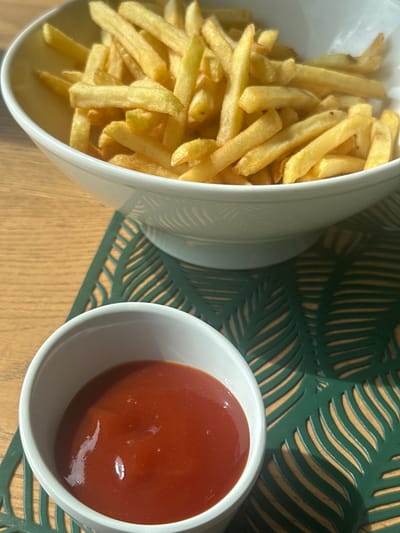 An overhead, close-up view captures a serving of French fries and a side of ketchup arranged on a table.
In the upper portion of the frame, a white ceramic bowl, approximately 20-25 cm in diameter, is filled with a substantial quantity of golden-brown French fries. The fries are cut uniformly, varying slightly in length and exhibiting a crispy texture. The bowl features a subtle, embossed or carved decorative pattern around its exterior, near the base.
Positioned below and slightly to the left of the large bowl, a smaller white ceramic ramekin, approximately 8-10 cm in diameter, holds a pool of viscous, reddish-orange ketchup. The surface of the ketchup reflects ambient light, indicating a smooth, liquid consistency.
Both the large bowl and the ramekin rest upon a dark green placemat. This placemat is adorned with an intricate, cut-out pattern resembling stylized palm leaves or tropical foliage, allowing the underlying surface to show through.
The objects are situated on a light brown wooden table surface, visible around the placemat. The wood grain is horizontally oriented. A distinct shadow from the larger bowl is cast onto the placemat and table, indicating a light source originating from the upper left of the frame. The scene is illuminated, suggesting an indoor setting.
No individuals are present in the image. The specific location within the city of Giebułtów, Poland, cannot be determined from the visual evidence provided in the image. The setting is consistent with a casual dining or home environment.