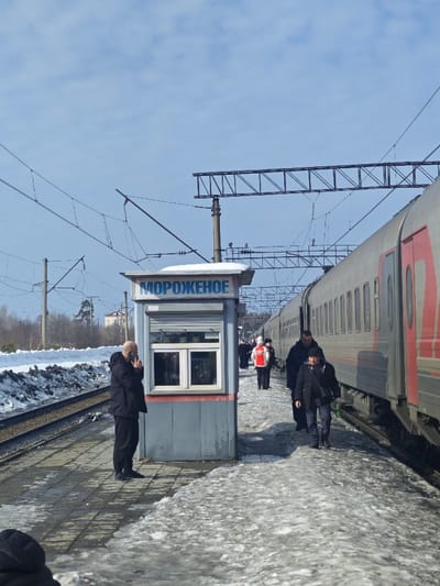 The image captures a train station platform in Druzhinino, Russia, during daylight under a bright, partly cloudy blue sky. A silver-grey passenger train with red accents is stopped at the platform on the right side of the frame. The platform is paved, with visible patches of melting snow and ice. Parallel to the platform, a set of railway tracks runs to the left, bordered by accumulated snow. Overhead electrical lines and supporting utility poles are present above the tracks and platform.
A small, grey kiosk with a white window frame and a closed roller shutter on its front is situated on the platform to the left of the train. The kiosk's roof has a layer of snow, and its sign displays "МОРОЖЕНОЕ" (ICE CREAM) in blue Cyrillic letters. A bald man in dark clothing stands to the left of the kiosk, facing right, holding a dark smartphone to his right ear as if engaged in a call.
On the platform to the right, adjacent to the train, several individuals are visible. Two men in dark jackets and pants walk away from the camera, along the platform next to the train. Further down the platform, a person wearing a red and white jacket stands facing left, with additional indistinct figures visible in the distance. The overall environment suggests a winter or early spring setting, with bare trees and some evergreens in the background.