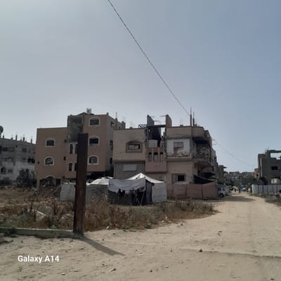 The image depicts a street in Nuseirat, Palestinian Territories. Damaged buildings are visible along the street. Some buildings are partially destroyed, with visible holes and structural damage. Tents or shelters are erected near the buildings. The road appears to be unpaved. A pole or post is visible in the foreground. The sky is clear.