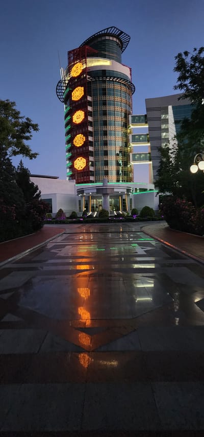 This is a vertical, low-angle shot of the magnificent "Türkmenistan" hotel in Türkmenbaşy, Turkmenistan, taken at dusk. The hotel is a striking architectural marvel, its cylindrical tower adorned with intricate patterns and glowing golden circular motifs that stand out against the twilight sky. The building's facade is a mosaic of glass and metal, reflecting the fading daylight and the colorful lights from within.

The foreground is dominated by a sleek, dark, reflective surface, likely a fountain or a polished plaza, which mirrors the building's illuminated features, creating a dazzling display of light and color. The reflections show abstract shapes and patterns of orange and green light. Lush green bushes and trees are visible around the base of the hotel, adding a touch of nature to the urban landscape. A modern lamppost with three glowing globes is also visible, adding to the ambient light.

The sky is a deep blue, indicating the transition from day to night. The overall mood is one of grandeur and modern elegance. There is no visible text in the image. The scene captures the impressive architecture of the hotel and its surroundings during a serene evening.