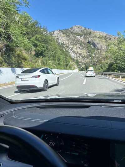 The image captures a view from inside a vehicle, looking forward through the windshield. The vehicle is on a paved road, likely a highway, winding through a mountainous and forested area. In the foreground, the dashboard of the vehicle is visible, with a digital display showing a speed of 76 km/h. A steering wheel is also partially visible on the left. On the road ahead, a white Tesla is driving in the same direction, slightly to the left. Further ahead, another white car is visible in the distance. The road is flanked by a low concrete barrier on the left and a metal guardrail on the right. The landscape is characterized by steep, rocky hills covered with dense green vegetation and trees, under a clear blue sky. The location is identified as Golubovići, Montenegro.