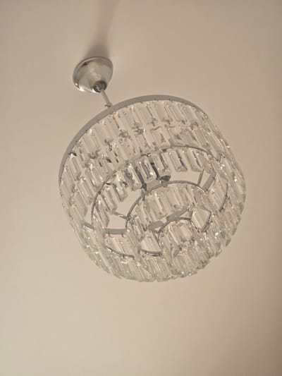 A close-up, low-angle shot shows a crystal chandelier hanging from a plain, off-white ceiling. The chandelier has a silver-toned metal frame and is adorned with multiple tiers of clear, rectangular crystal prisms. The lighting in the image is soft, casting a gentle shadow on the ceiling to the left of the chandelier. The image is a still photograph with no people, actions, or interactions. The specific location in Avtury, Russia, is not depicted or discernible from the visual information provided in the image.