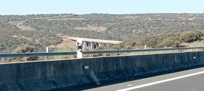The image shows a highway overpass in Jaraicejo, Spain. The overpass is a concrete structure with multiple pillars supporting a roadway above a wooded hillside. A red vehicle is visible on the road of the overpass. In the foreground, there is a concrete barrier and a metal guardrail, typical of highway infrastructure. The background consists of a hilly landscape covered in dense green vegetation, likely trees. The sky is clear and blue, indicating daytime.