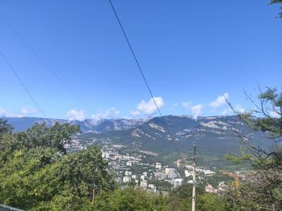 The image captures a panoramic view of a coastal city nestled at the foot of a mountain range under a clear blue sky. Several power lines traverse the upper portion of the frame. In the foreground, lush green trees and foliage obscure parts of the scene. Below the trees, the city unfolds with a dense cluster of white and light-colored buildings of varying heights, suggesting residential and commercial areas. The buildings are interspersed with greenery, indicating a well-treed urban environment.

Further in the distance, the imposing mountain range rises, its slopes covered in dark green vegetation, with rocky outcrops and lighter-colored geological formations visible along the peaks. A few scattered clouds drift over the mountain tops. A prominent yellow construction crane is visible within the city, hinting at ongoing development or infrastructure projects. The overall impression is one of a vibrant coastal settlement situated in a picturesque natural setting. Based on the visual cues, this scene is likely located in the vicinity of Massandrovsky village council, Ukraine, given the characteristic mountainous terrain and coastal urban development often found in the Crimean peninsula.