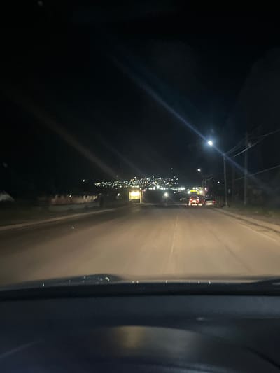 The image depicts a nighttime view from inside a vehicle driving on a road, possibly in the city of Arsha, Georgia. The road surface is visible in the foreground and extends into the distance. Streetlights illuminate the scene, casting a warm glow on the asphalt. In the middle ground, there are several vehicles, including cars and what appears to be a larger vehicle, possibly a truck or bus, with its lights on. The background shows a town or city with numerous lights scattered across hillsides, suggesting a populated area. The sky is dark, with some faint light streaks, possibly from streetlights or distant lights reflecting off clouds. The overall impression is that of a drive through a town at night.