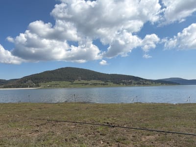 The image shows a scenic view of Batak Reservoir near Rakitovo, Bulgaria. The foreground features a grassy bank with fishing rods laid out. The water of the reservoir extends to the middle ground, with a forested hill in the background. The sky is blue with fluffy white clouds.