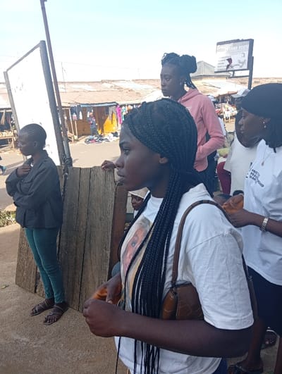 The image depicts a street scene in Jebbu Miango, Nigeria, on a clear day. In the foreground, five individuals, primarily young women, are visible. The closest figure, a young woman with long dark braids, stands in profile facing right, wearing a white short-sleeved t-shirt featuring a large photographic print of a light-skinned person's face. She carries a dark brown, textured shoulder bag on her right side, and her left hand is partially clenched. To her right and slightly behind, another young woman, also facing right, wears a white t-shirt with "PRISTINE PRAISE WORSHIP" text, and has a dark head covering or bunched hair. A silver bracelet is on her right wrist. Further back, a young woman in a light pink hooded sweatshirt with dark hair tied up looks to the right. To the far left, a young woman with short dark hair, wearing a dark jacket and teal pants, stands with crossed arms, looking left. Partially visible between the central figures is a man in a white cap.
The background shows a bustling street market environment with numerous low-rise structures, likely shops or stalls, featuring corrugated metal roofs. Merchandise, including clothing, is displayed outside these establishments, and items are visible on the ground along the street. A large black sign with "BREAKTHROUGH FASHION HOUSE" and other white text and graphics is mounted above the background buildings on the right. To the left, a large, blank white signboard is affixed to a pole. The ground is an uneven mix of dirt and possibly concrete. A wooden plank barrier separates the immediate foreground from the street and market stalls. The sky above is clear and light blue.