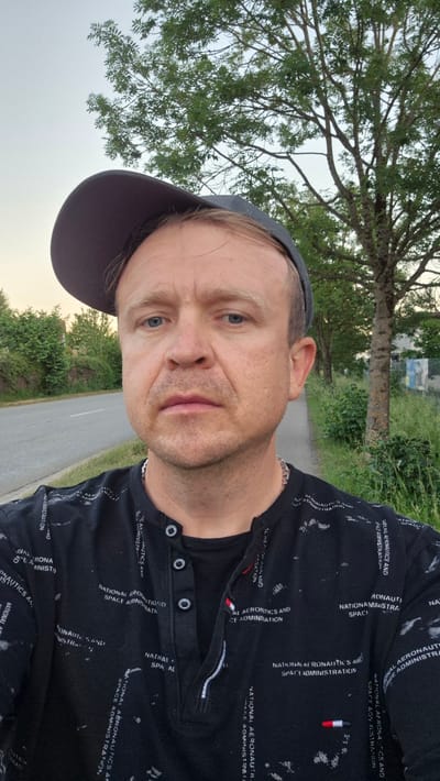 A medium-shot photograph depicts a middle-aged man wearing a black baseball cap and a black shirt with printed white lettering of "NATIONAL AERONAUTICS AND SPACE ADMINISTRATION." He appears outdoors near Essenbach, Germany, with a tree to his right, a grassy area and road visible behind him. He is facing the camera with a neutral expression.