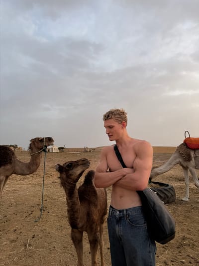 In Douar Ait Daoud, Morocco, under a cloudy sky, a shirtless man with blonde curly hair stands with his arms crossed. He is wearing blue denim jeans and a black bag is slung across his body. To his left, two camels stand in a dusty, dry terrain. One camel, larger than the other, is tethered by a blue rope. To the man's right, another camel is visible, equipped with an orange saddle. In the background, a few tents and structures suggest a camp or village.